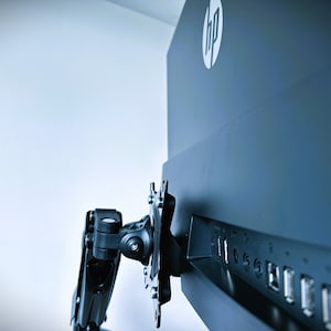 Könnte beinhalten: Ein schwarzer HP-Computermonitor, montiert an einem schwarzen, verstellbaren Arm. Das HP-Logo ist weiß. Die Rückseite des Monitors zeigt USB-Anschlüsse und andere Anschlüsse.