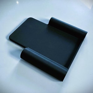 May include: A black plastic phone stand with a curved design. The stand is designed to hold a phone upright.