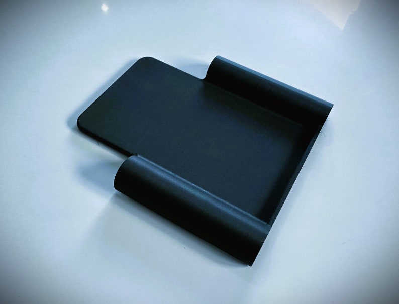 May include: A black plastic phone stand with a curved design. The stand is designed to hold a phone upright.