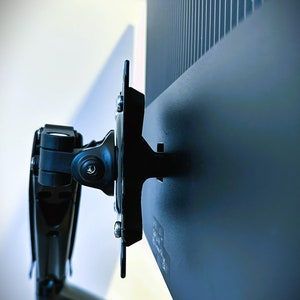 May include: A black monitor arm with a metal arm and a black plastic base. The arm is attached to the back of a black monitor.
