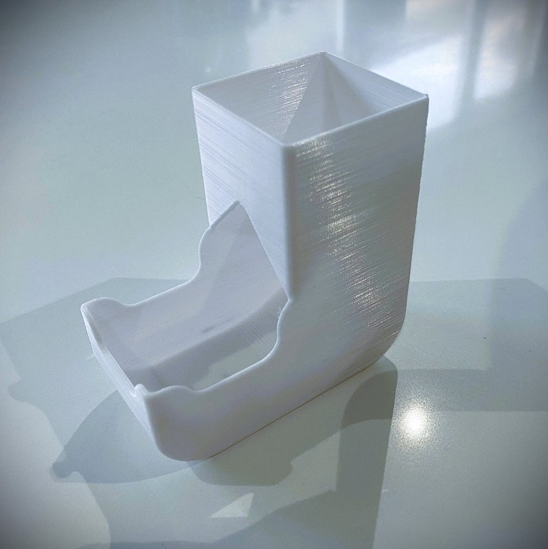 May include: White plastic dice tower with a square top and a curved bottom. The tower is designed to hold dice and release them through a chute.