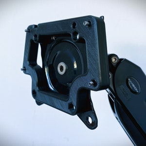 Op de afbeelding: Een zwarte 3D-geprinte beugel voor een monitorarm, met meerdere schroefgaten en een ronde opening. De beugel is bevestigd aan een zwarte monitorarm met een zilveren knop met het opschrift "InVision".