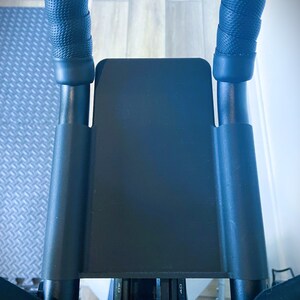 May include: Black exercise bike handlebars with black foam grips and a black metal frame. The handlebars have a black plastic cover with a red and black digital display that reads "10 20 30 40 50 60 70".