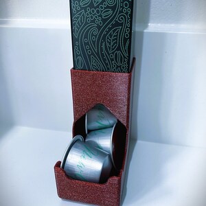 May include: A red plastic holder with a black box on top. The holder has a slot for holding coffee pods. There are four silver coffee pods inside the holder.