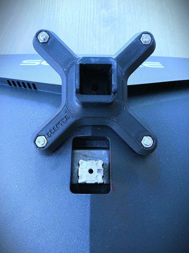Pode incluir: Um suporte de monitor de pl&aacute;stico preto com quatro furos para parafusos e uma abertura quadrada no centro. O suporte &eacute; projetado para ser fixado na parte de tr&aacute;s de um monitor.