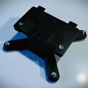 May include: Black plastic phone mount with four legs and multiple screw holes. The mount is designed to hold a phone securely in place.