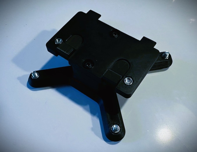 May include: Black plastic phone mount with four legs and multiple screw holes. The mount is designed to hold a phone securely in place.