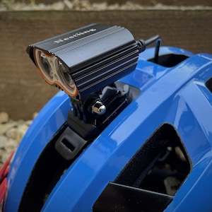 Könnte beinhalten: Ein schwarzes Fahrradhelmlicht mit zwei hellen weißen LED-Leuchten und dem Wort "Nesting" auf der Oberseite. Das Licht ist an einem blauen Fahrradhelm montiert.