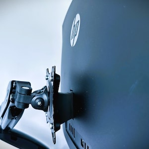 Puede incluir: Un brazo de monitor negro con un brazo giratorio y una placa de montaje unida a la parte posterior de un monitor HP negro.
