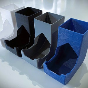 May include: Four dice towers in different colors: white, black, gray, and blue. The towers are made of plastic and have a textured surface.