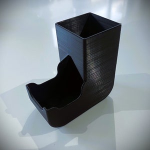 May include: A black plastic dice tower with a square top and a curved bottom. The tower is designed to hold dice and allow them to roll out of the bottom.