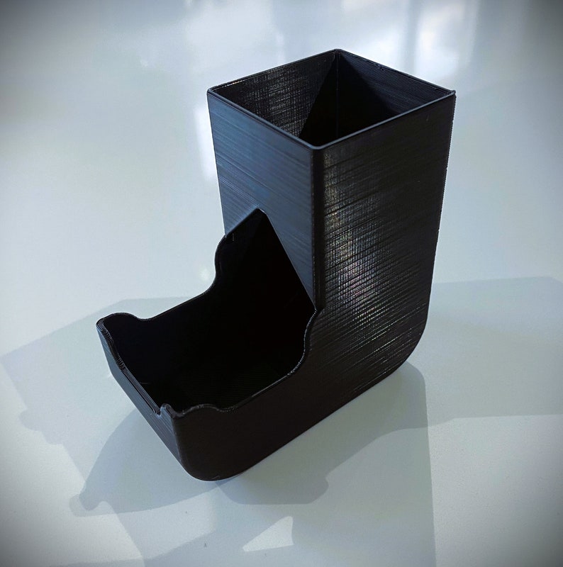 May include: A black plastic dice tower with a square top and a curved bottom. The tower is designed to hold dice and allow them to roll out of the bottom.