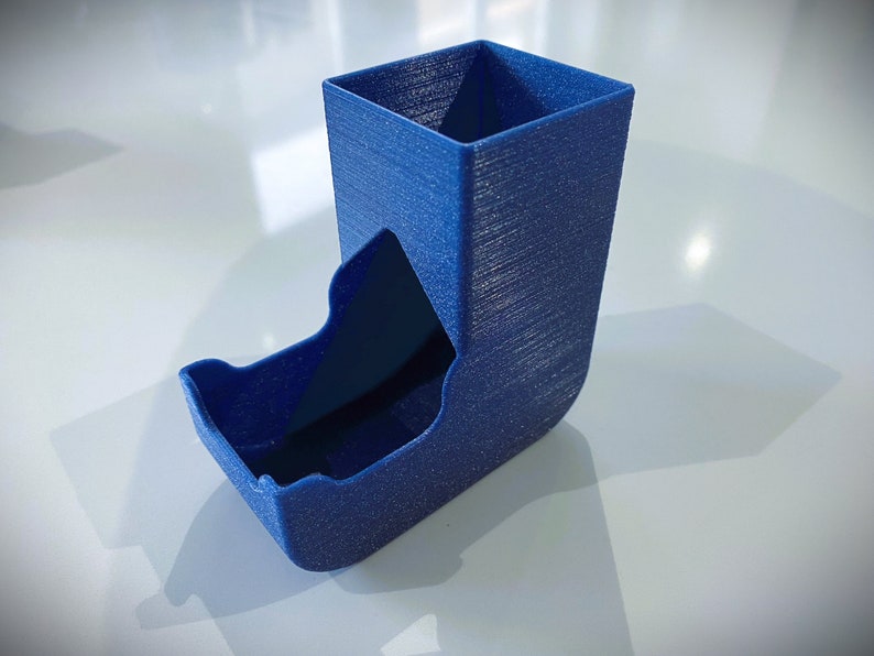May include: A blue 3D printed dice tower with a square top and a curved bottom. The tower has a textured surface and a slot for dice to roll out.