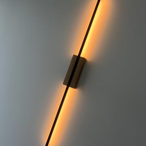 May include: A modern wall sconce with a long, thin, black light bar mounted on a wooden base. The light bar emits a warm, yellow glow.