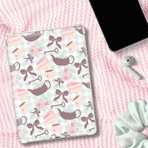 Puede incluir: Una funda para tableta con un patrón caprichoso que presenta teteras, rebanadas de pastel, lazos, llaves y tazas de té sobre un fondo a cuadros verde claro y blanco. La funda es transparente y tiene un diseño protector.