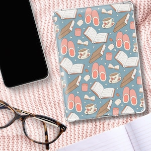 Peut inclure: Une coque de tablette avec un fond bleu clair orné d'un motif répété de livres, tasses à thé, pantoufles, bougies et nœuds. L'étui est entouré d'une couverture en tricot rose, avec une paire de lunettes et un smartphone dans le cadre.