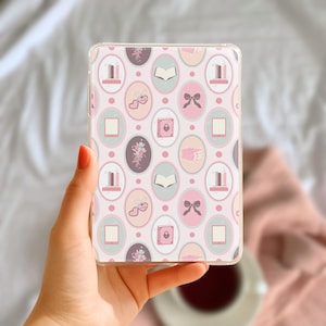 Puede incluir: Una funda transparente para teléfono con un patrón repetido de formas ovaladas en rosa, gris y verde menta. Los óvalos contienen ilustraciones de libros, lazos, corazones y flores. La funda se sostiene en una mano con esmalte de uñas rojo.