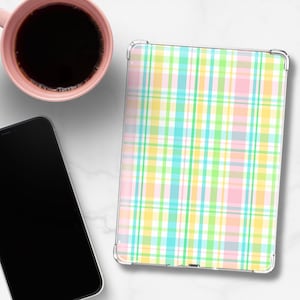 May include: A tablet case with a pastel plaid pattern in pink, yellow, green, and blue. A pink mug with dark liquid and a black smartphone are also visible on a white surface.