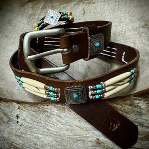 Peut inclure: Une ceinture en cuir marron avec des accents argentés et turquoise. La ceinture est ornée d'un motif en os et en perles et d'une boucle argentée.