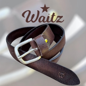 Op de afbeelding: Een bruine leren riem met een zilveren gesp. De riem heeft een versleten uiterlijk en een klein, in reliëf aangebracht logo bij de punt. Het woord "Waitz" wordt in een scriptlettertype boven de riem weergegeven.