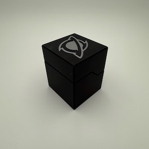 May include: A black square box with a silver emblem on the top. The box is closed and sits on a white surface. The emblem is a stylized design with curved lines.