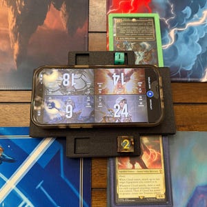 May include: A black phone displaying game information rests on a black stand, with dice and game cards nearby. The phone shows numbers 18, 9, 14, and 24. The scene is set on a gaming mat with colorful graphics.