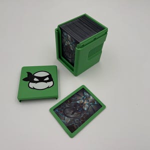May include: Green card holder with a black and white cartoon character design. The holder is open, revealing a stack of cards. Two additional cards are displayed, one with the character design and the other with a graphic image.