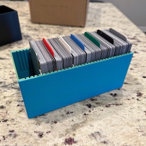 May include: A turquoise blue card holder filled with a stack of cards. The cards are separated by colored dividers: red, white, blue, and green. A black box and a cardboard box are in the background.