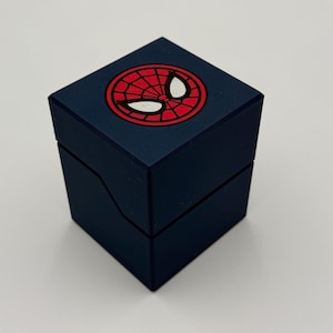 May include: A dark blue, square box with a hinged lid. The top of the box features a red and white Spider-Man logo. The box is likely for jewelry or a small gift.