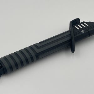 May include: A black lightsaber with a textured grip and a silver button. The lightsaber has a black and silver handle with a silver button on the side.