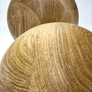 May include: Two spherical wooden objects with a light brown color and visible wood grain. The objects are positioned one on top of the other, with the top one partially visible. The wood grain creates a textured surface.