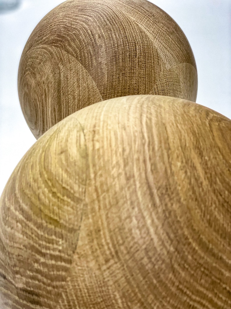 May include: Two spherical wooden objects with a light brown color and visible wood grain. The objects are positioned one on top of the other, with the top one partially visible. The wood grain creates a textured surface.