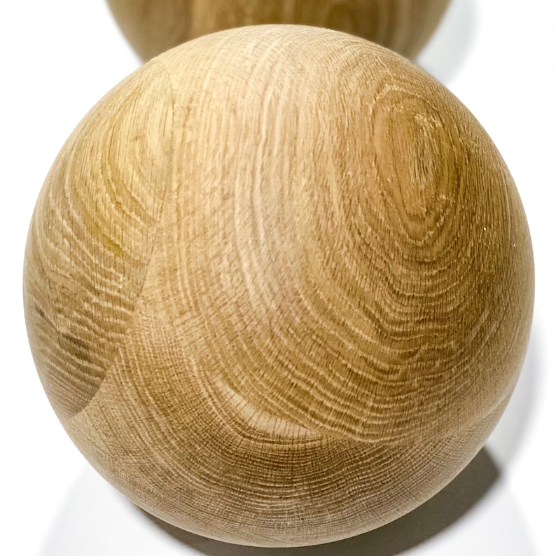 May include: Two wooden spheres with a natural wood grain pattern. The spheres are light brown and appear to be made of oak or a similar hardwood. The wood grain creates a swirling pattern across the surface of the spheres.