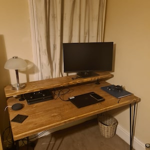 Puede incluir: Un escritorio de madera con un marco de metal negro y un monitor negro en un estante. El escritorio tiene una tapa de madera marrón y una lámpara negra en el lado izquierdo. Hay una computadora portátil y un teléfono en el escritorio.