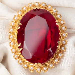 Può includere: Spilla dorata con una grande gemma rossa ovale sfaccettata. La gemma è circondata da piccole perle bianche e da una cornice dorata decorativa. La spilla è posta su uno sfondo di tessuto bianco. La spilla misura circa 5 cm di lunghezza.
