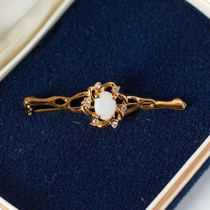 Könnte beinhalten: Goldfarbene Brosche mit einem ovalen, weißen Mittelstein, umgeben von kleinen, klaren Kristallen. Die Brosche hat ein dekoratives, offenes Design. Sie wird in einer blau samtgefütterten Schatulle mit cremefarbenem Innenraum präsentiert.