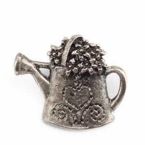 May include: A silver-colored watering can-shaped pin. The watering can is filled with flowers and has a heart-shaped design on the front. The pin has a handle and a spout.