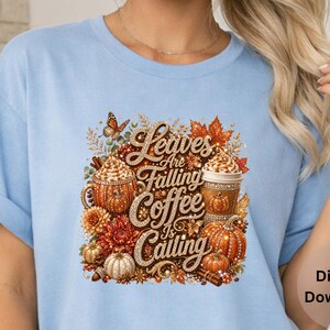 May include: A light blue t-shirt featuring a graphic print with the text Leaves Are Falling Coffee Is Calling, surrounded by illustrations of pumpkins, autumn leaves, coffee cups with whipped cream, a butterfly, and cinnamon sticks.