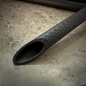 May include: Close-up of a black carbon fiber tube with a textured, woven pattern. The tube has a rounded, oval-shaped opening at the end. The background is a mottled, neutral-toned surface.