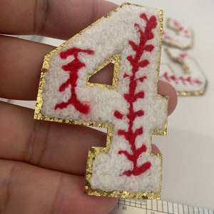 Pode incluir: Um patch de feltro branco com o número 4 e costuras vermelhas, que se assemelha a uma bola de beisebol. O patch tem uma borda dourada com glitter. Patches adicionais são visíveis no fundo.