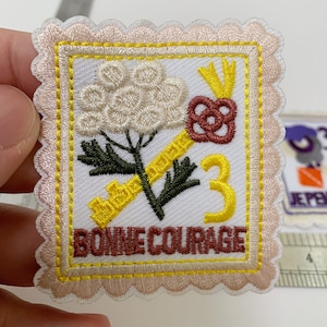 May include: Embroidered iron-on patch featuring a white flower, a gold key, and a brown heart. The patch has a yellow border and the words "BONNE COURAGE" in brown text.