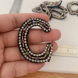 以下が含まれることがあります： 金色のビーズとカラフルなラインストーンで飾られた黒いフェルトの文字C。文字の高さは約5cmです。
