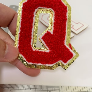 Könnte beinhalten: Roter Filzbuchstabe Q mit goldfarbenem Glitzerumrandung. Der Buchstabe ist etwa 5 cm hoch.