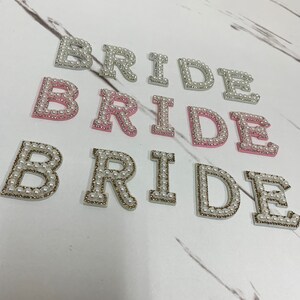 May include: Three sets of the word "BRIDE" spelled out in pearl-embellished letters. The first set is white, the second is pink, and the third is white with gold accents.