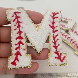 May include: White felt letter M with red stitching, resembling a baseball, outlined in gold glitter. The letter is approximately 1 inch tall.