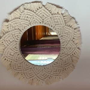 May include: A round mirror with a white macrame frame. The mirror is reflecting a room with wooden floors and purple curtains.