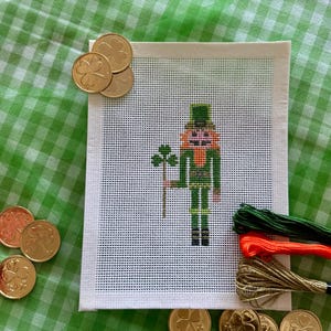 May include: A cross-stitch of a leprechaun holding a shamrock, with gold coins and embroidery floss. The leprechaun is wearing a green hat and suit. The background is white with a green grid pattern.