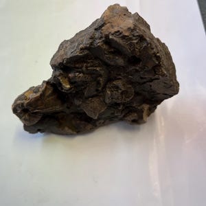 Könnte beinhalten: Ein dunkelbrauner, unregelmäßig geformter Felsen mit einer rauen, strukturierten Oberfläche. Der Felsen hat ein natürliches, verwittertes Aussehen mit verschiedenen Vorsprüngen und Vertiefungen. Die Farbe ist eine Mischung aus Dunkelbraun und Hellbraun, was auf Mineralablagerungen hindeutet.