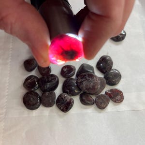 Könnte beinhalten: Eine Sammlung dunkelroter Edelsteine, einige mit glänzender Oberfläche, auf einer weißen Fläche. Ein Edelstein wird von einer Hand hochgehalten und zeigt ein leuchtend rotes Inneres.