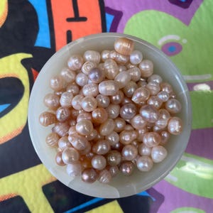 Könnte beinhalten: Draufsicht auf eine kleine, cremefarbene Schale, gefüllt mit zahlreichen Süßwasserperlen. Die Perlen variieren in der Farbe, mit Schattierungen von Rosa, Pfirsich und Creme. Die Schale steht auf einer bunten, gemusterten Oberfläche.